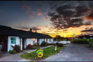 Campingplatz mit kleinen Häusern, gepflegtem Garten und Kiesweg, im Hintergrund Autos und ein Sonnenuntergang am Himmel.