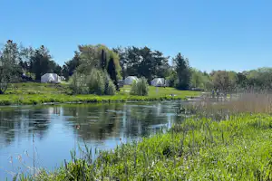 Campingplatz am See mit grünem Ufer, Schilf und Bäumen; im Hintergrund Zelte und kleine Gebäude am Wasser.
