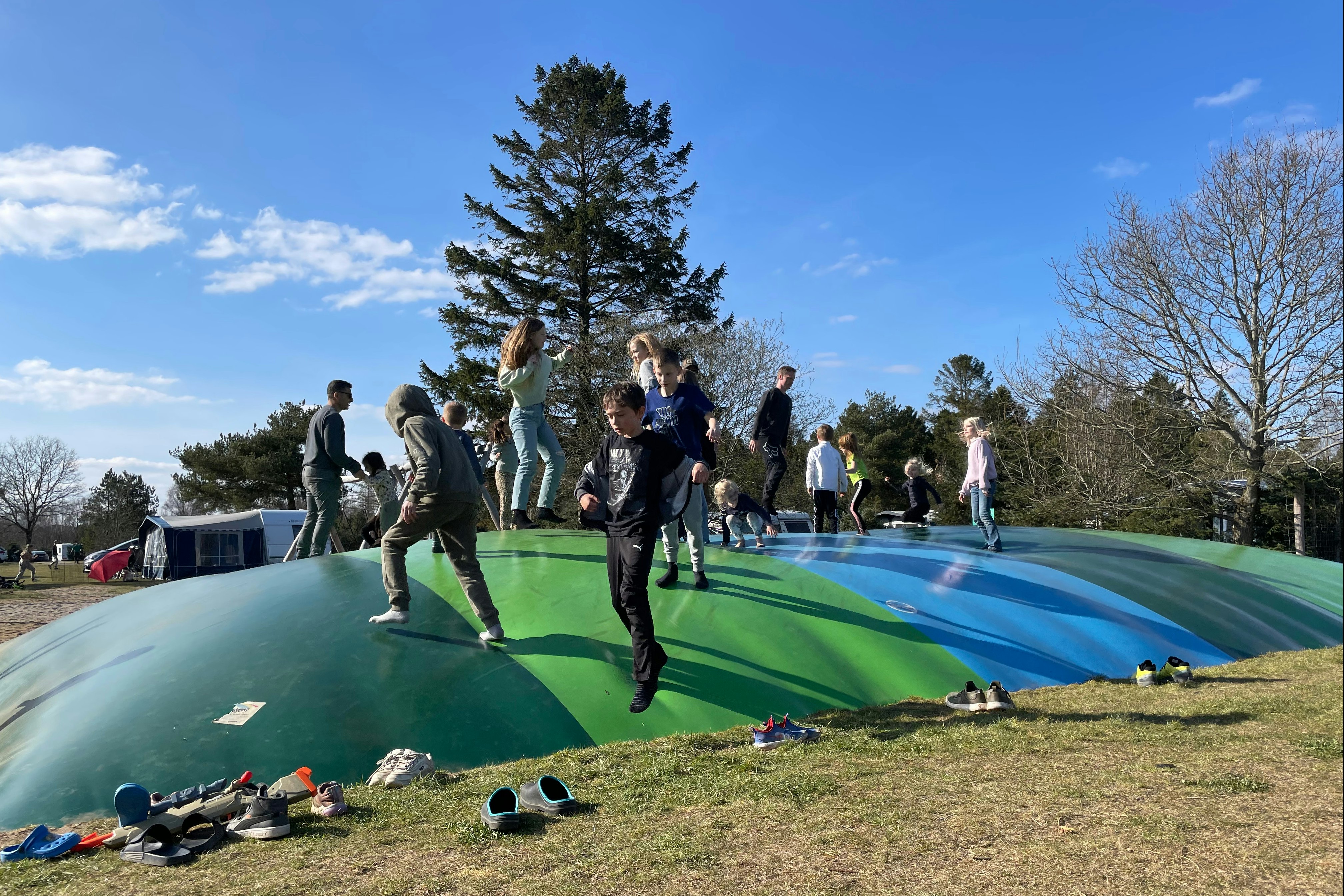 Gruppe springt auf einem Trampolin auf dem Campingplatz, im Hintergrund Bäume und ein blauer Himmel.