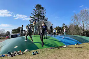 Gruppe springt auf einem Trampolin auf dem Campingplatz, im Hintergrund Bäume und ein blauer Himmel.