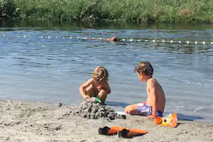 Zwei Kinder sitzen am Seeufer im Sand, spielen mit Wasserspielzeug und liegen am Strand.