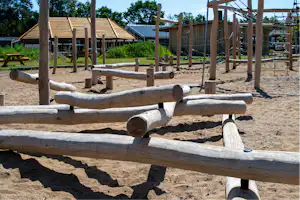 Holzbalancierbalken auf einem Camping-Spielplatz, im Hintergrund ein Holzhaus und ein Zaun mit Bäumen.