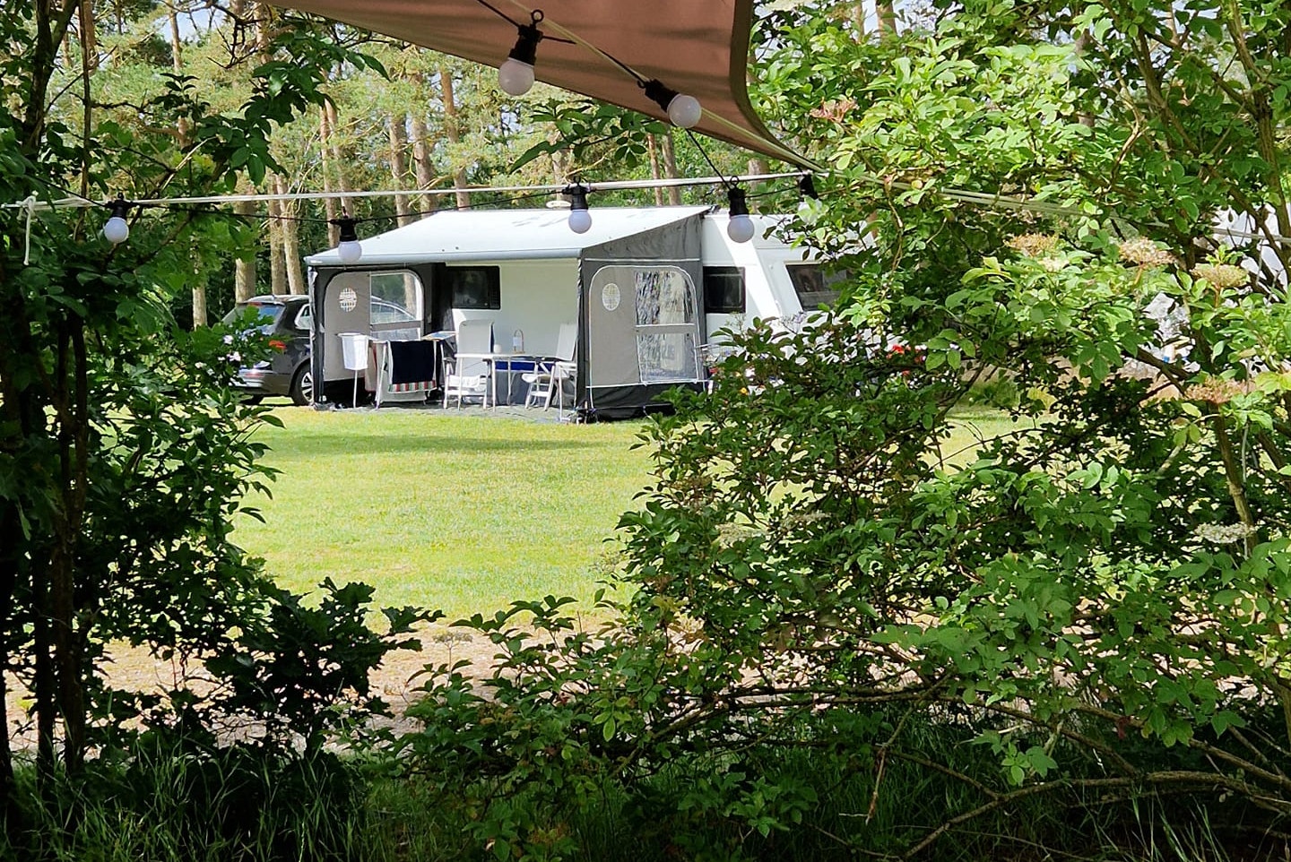 Wohnwagen mit Vorzelt und Sitzbereich auf einem Campingplatz im Grünen, umrahmt von Bäumen und Rasen.