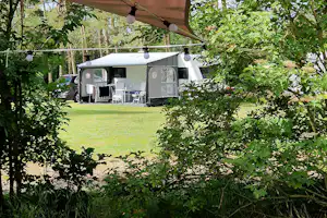 Wohnwagen mit Vorzelt und Sitzbereich auf einem Campingplatz im Grünen, umrahmt von Bäumen und Rasen.