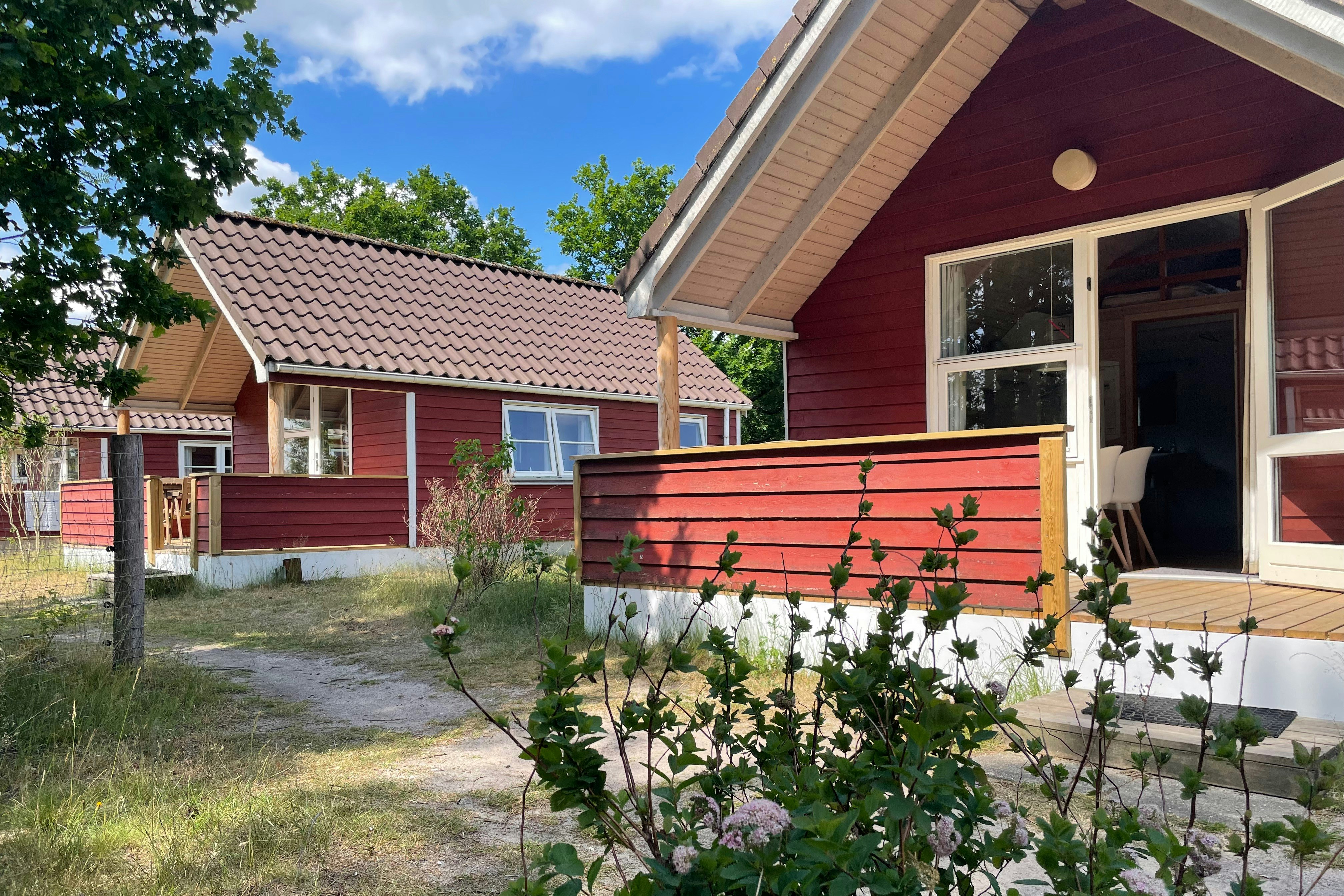 Rotes Ferienhaus mit Terrasse und großen Fenstern, davor ein Weg und ein Garten mit blühenden Sträuchern.