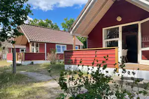 Rotes Ferienhaus mit Terrasse und großen Fenstern, davor ein Weg und ein Garten mit blühenden Sträuchern.