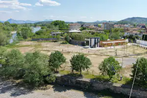 Blick auf einen Campingplatz mit eingezäuntem Gelände, Bäumen, Wegen und mehreren Gebäuden in einer hügeligen Landschaft.