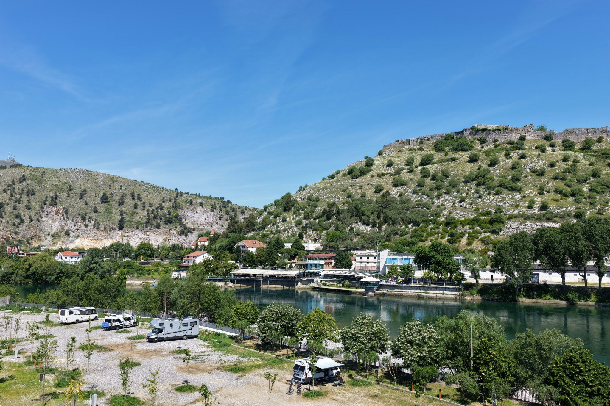 Campingplatz am See mit Booten am Steg, Wohnwagen und Zelten auf dem Platz, Gebäude am Ufer und Hügel im Hintergrund.