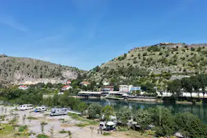 Campingplatz am See mit Booten am Steg, Wohnwagen und Zelten auf dem Platz, Gebäude am Ufer und Hügel im Hintergrund.