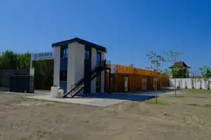 Kleines Campinghaus mit Treppe, Zaun und Holzverkleidung, daneben Kiesweg und Rasenfläche vor blauem Himmel.
