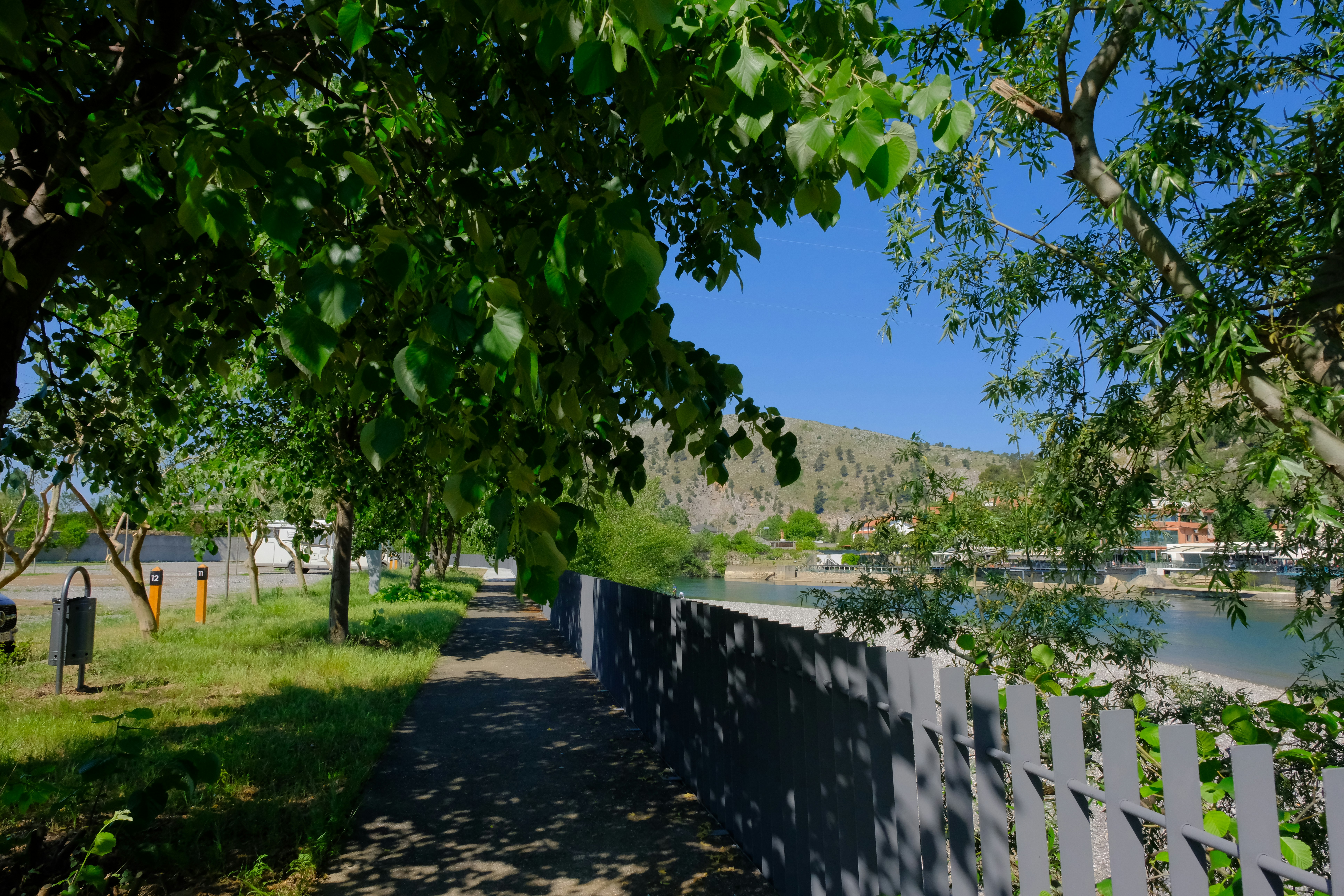 Campingweg mit Holzlattenzaun, Bäumen und Rasenfläche, im Hintergrund Häuser und Berge unter blauem Himmel.