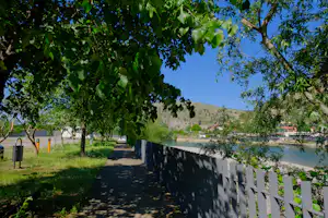 Campingweg mit Holzlattenzaun, Bäumen und Rasenfläche, im Hintergrund Häuser und Berge unter blauem Himmel.