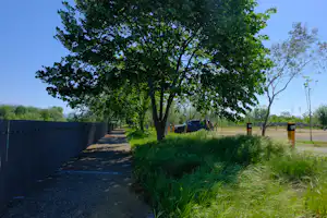 Campingweg entlang eines Zauns, daneben Gras und Büsche, im Hintergrund Bäume und ein Zeltbereich.