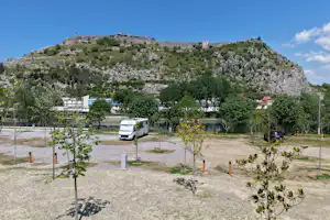 Campingplatz mit Wohnmobil auf Kiesstellplatz, jungen Bäumen und Blick auf einen bewaldeten Berg im Hintergrund.