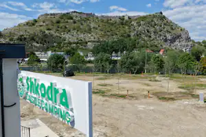 Eingangsschild am Campingplatz mit Schriftzug „Castelcamping“ vor einem gepflegten Gelände und Bäumen, im Hintergrund ein bewachsener Hügel.