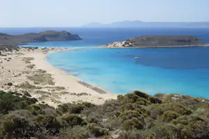 Türkisfarbene Bucht mit Sandstrand, mehreren Segelbooten im Wasser und Dünenlandschaft vor Hügeln und Meer.