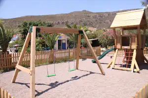 Spielplatz am Campingplatz: Holz-Klettergerüst mit Rutsche, Schaukeln und Sandboden, im Hintergrund Palmen und Berge.