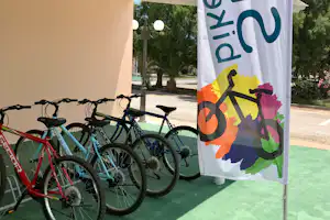 Mehrere Fahrräder stehen vor einer weißen Wand und unter einem bunten Werbebanner, im Campingbereich mit Bäumen im Hintergrund.