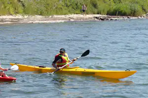 Zwei Kajaks auf dem Wasser, eine Person paddelt im gelben Kajak, im Hintergrund Ufer mit Bäumen und Kiesstrand.