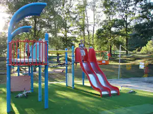 Spielplatz am Campingplatz mit blauer Rutsche, roten Schaukeln und eingezäuntem Bereich auf grünem Rasen zwischen Bäumen.