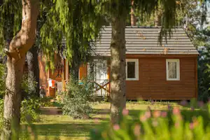 Holzhaus auf einem Campingplatz zwischen Bäumen, mit Veranda und Stufen, im Vordergrund grünes Gras.
