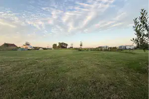 Weite Wiese auf einem Campingplatz mit Wohnwagen und Zelten in der Ferne, Bäume und ein Weg am Rand.