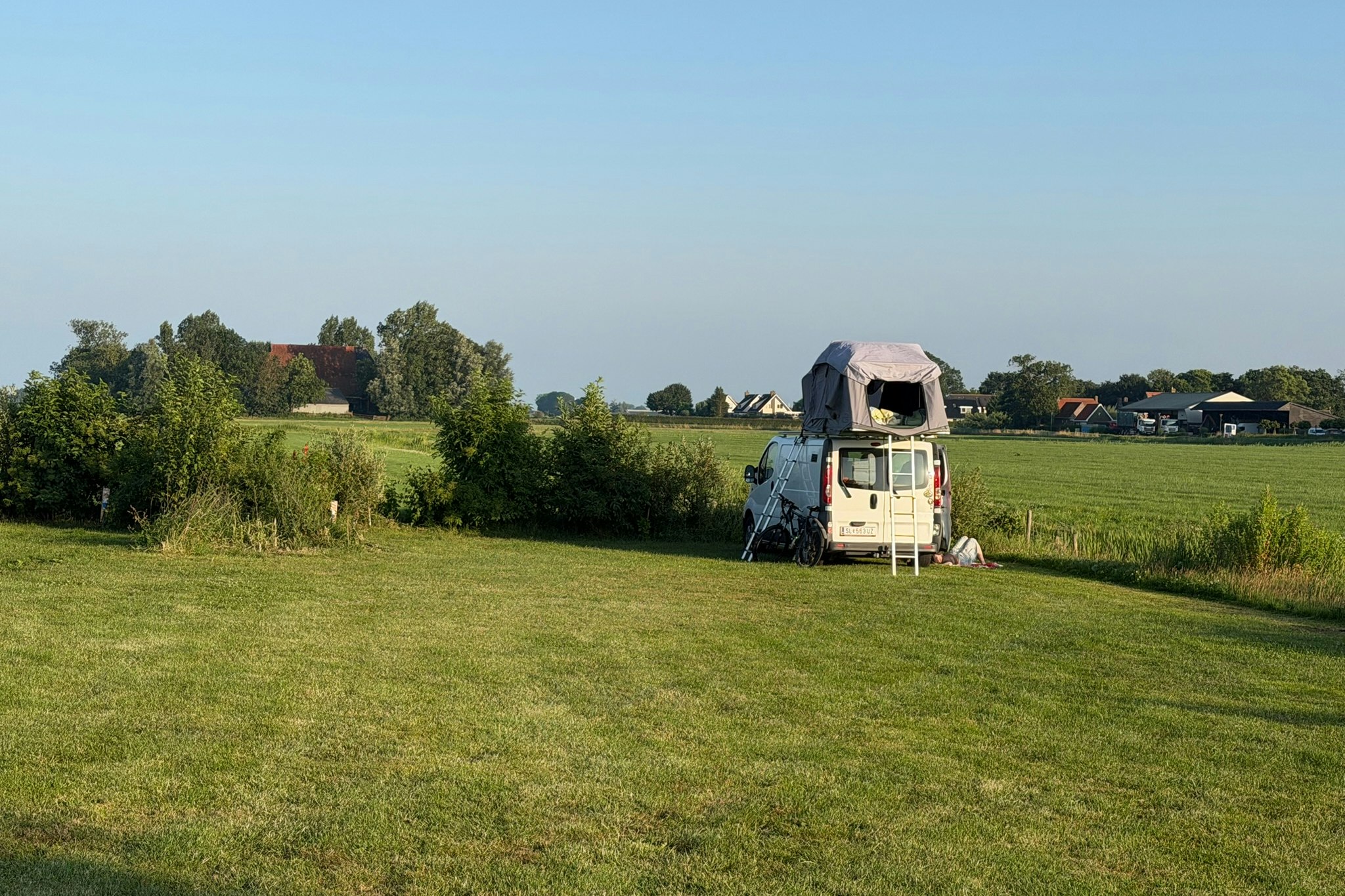 Wohnwagen mit Vorzelt auf einem Campingplatz, umgeben von grünen Wiesen und Bäumen, im Hintergrund Häuser und Felder.