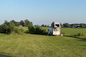 Wohnwagen mit Vorzelt auf einem Campingplatz, umgeben von grünen Wiesen und Bäumen, im Hintergrund Häuser und Felder.