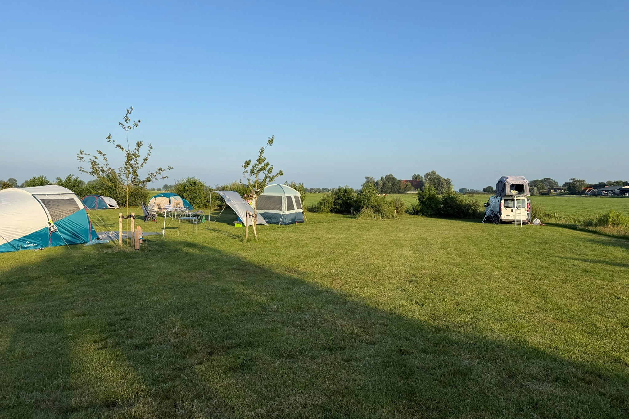 Campingplatz mit mehreren Zelten und Wohnmobilen auf einer grünen Wiese, Bäume und ein Weg im Hintergrund.