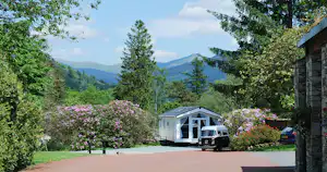 Wohnwagen und Auto vor einem kleinen Gebäude auf einem Campingplatz, umgeben von Bäumen, Blumen und Blick auf Berge.