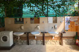 Camping-Badezimmer mit drei Waschbecken, Wandarmaturen und Spendern, unter Bäumen mit Blick auf ein Holzgeländer.