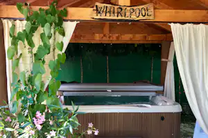 Whirlpool auf einer Terrasse unter Holzvordach, mit weißen Vorhängen, Pflanzen und Holzbalken im Hintergrund.