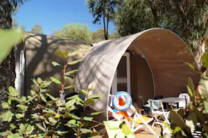 Campingzelt im Grünen mit Sonnenschirm, Liegestühlen und Tisch vor dem Zelt, umgeben von Bäumen und Sträuchern.