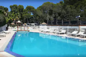 Blauer Campingpool mit Liegen, Sonnenschirmen und Holzterrasse, umgeben von Zäunen und Bäumen im Hintergrund.