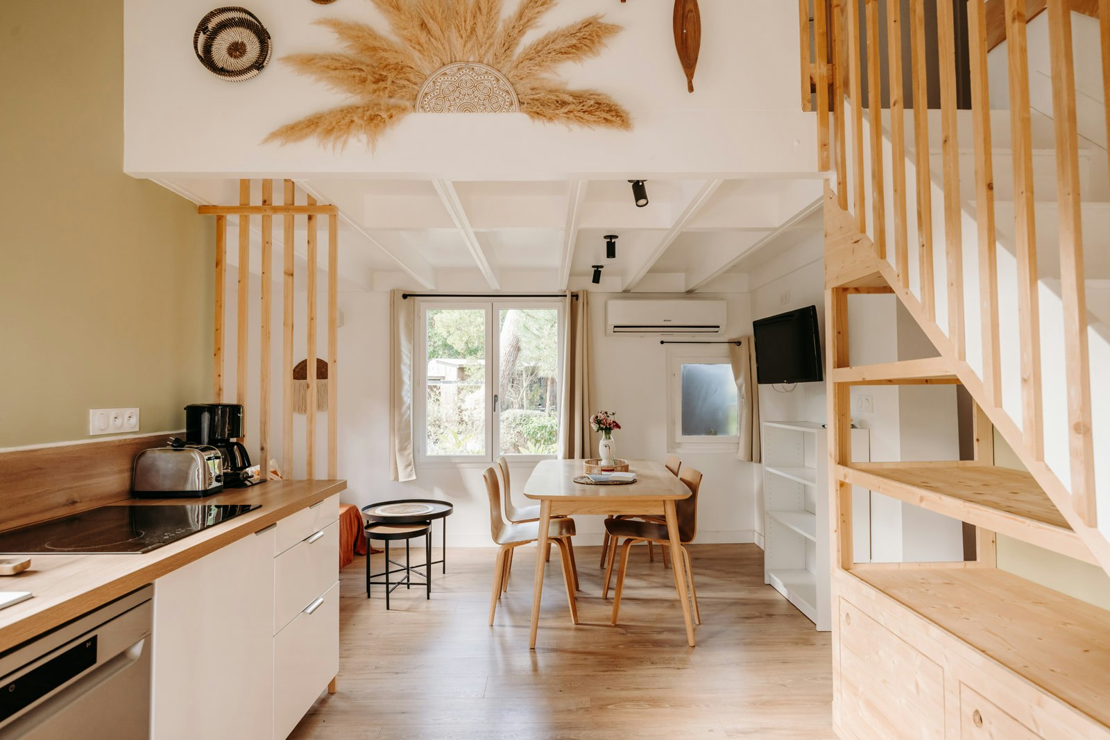 Innenraum mit Küchenzeile, Esstisch und Stühlen sowie Treppe zum Schlafbereich, große Fenster und Holzregale im Wohnbereich.