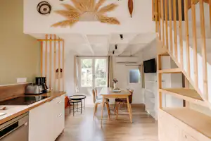 Innenraum mit Küchenzeile, Esstisch und Stühlen sowie Treppe zum Schlafbereich, große Fenster und Holzregale im Wohnbereich.