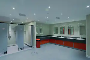Helles Sanitärgebäude mit Waschbecken, Spiegeln und beleuchteten Spendern, daneben Kabinen mit Türen und Handtuchhaken.