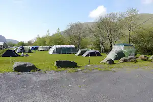 Campingplatz mit mehreren Zelten auf einer Wiese, Steinen im Vordergrund und Bäumen am Rand, im Hintergrund grüne Hügel.