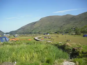 Campingplatz im grünen Tal mit Zelten, Weg und Sitzbank, im Hintergrund Berge und ein kleines Gewässer.