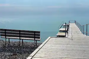 Holzsteg am See mit Geländer und Treppe, im Vordergrund ein Steg mit Bank und Kiesufer.