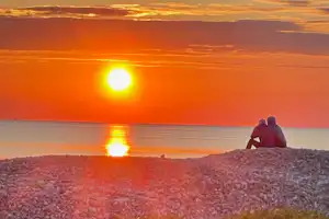 Sonnenuntergang am Meer: Ein Paar sitzt auf einem Hügel mit Blick auf die spiegelnde Wasserfläche.