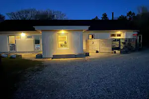 Beleuchteter Camping-Bungalow am Abend mit Kiesweg, Sitzbank und mehreren Fenstern, davor eine kleine Terrasse und Mauerwerk.