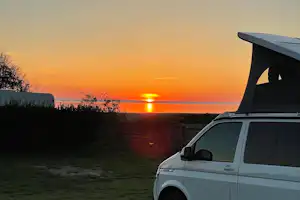 Weißer Camping-Van am Ufer mit Blick auf den Sonnenuntergang über dem See, im Hintergrund Bäume und Gras.