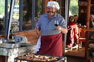 Mann am Grill mit Weinglas, Kochmütze und kariertem Hemd auf der Holzterrasse vor dem Campinghaus.