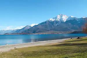 Ufer am See mit Kiesstrand, grüner Wiese und Blick auf hohe Berge in der Ferne.