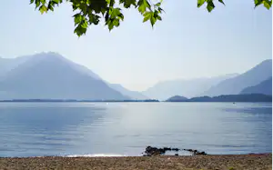 Seeufer mit Kiesstrand, ruhigem Wasser und Bergpanorama in der Ferne, im Vordergrund Blätter am Bildrand.