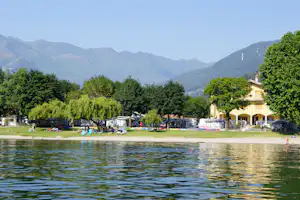 Blick auf den See mit Campingplätzen am Ufer, Wohnwagen und Zelte, Bäume und Berge im Hintergrund.
