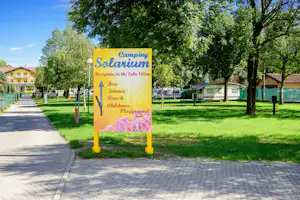 Schild am Campingplatz Solarium mit Hinweisen zu Bad, Beach und Wellness, daneben gepflasterter Weg und Rasen mit Bäumen.