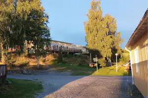 Campingweg zwischen Gebäuden, Bäume und Kiesfläche, rechts ein Haus mit Veranda, im Hintergrund weitere Gebäude.