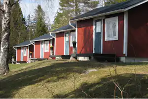 Rote Camping-Bungalows mit weißen Fenstern und Veranda, davor Rasen und Gehweg, im Hintergrund weitere Bungalows und Bäume.